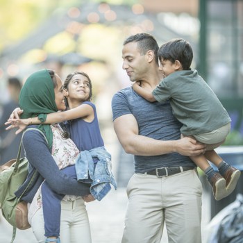 A cute little muslim family smiles at each other. The parents are carrying their kids and the kids have their arms wrapped around their parents.